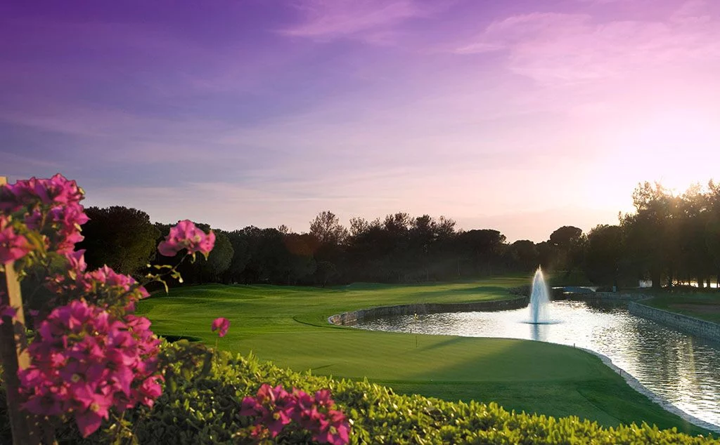 A stunning golf course in Belek, Antalya, with a fountain lake, lush greens, and a vibrant sunset sky.