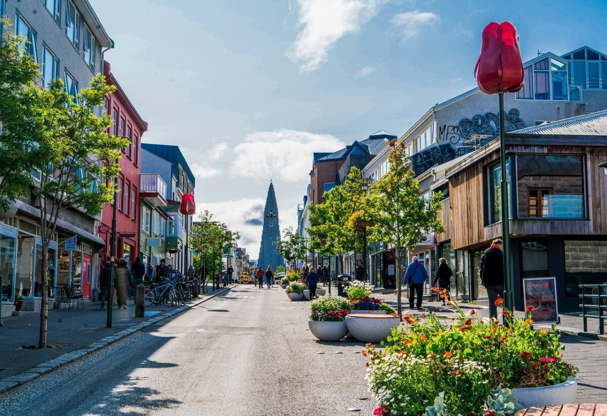 A sunny street scene in central Reykjavik, Iceland, with colourful low-rise buildings lining a pedestrian-friendly road, flowering plants and red tulip sculptures in the foreground, people walking casually, and the distinctive spire of Hallgrimskirkja church visible at the end of the street under a blue sky with light cloud.