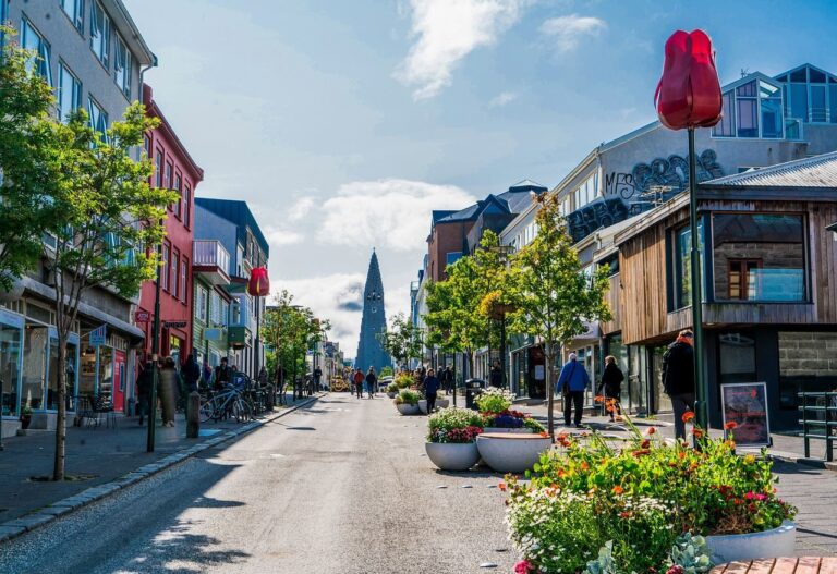 Tours 10 A sunny street scene in central Reykjavik, Iceland, with colourful low-rise buildings lining a pedestrian-friendly road, flowering plants and red tulip sculptures in the foreground, people walking casually, and the distinctive spire of Hallgrimskirkja church visible at the end of the street under a blue sky with light cloud.