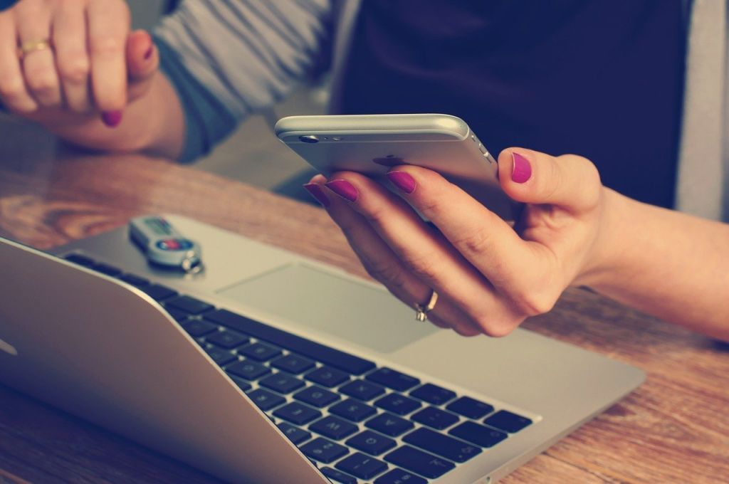 Close-up of a person using a smartphone while working on a laptop at a desk.