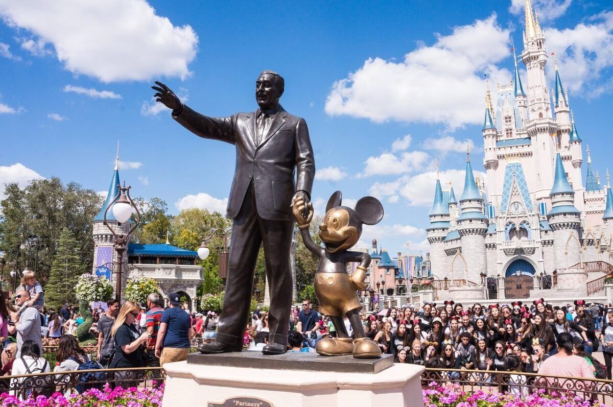 The Partners Statue of Walt Disney and Mickey Mouse in front of Cinderella Castle at Magic Kingdom, with a large crowd of visitors and bright blue skies.