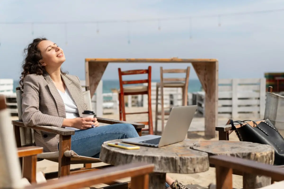 Digital nomad woman sitting outdoors at a beachside cafe with a laptop on a wooden table, enjoying the sun with her eyes closed and holding a cup of coffee.