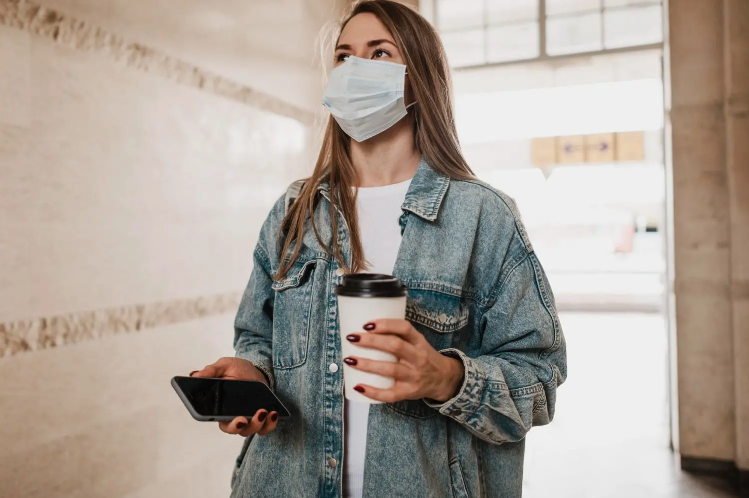 woman wears face mask and holds phone and coffee