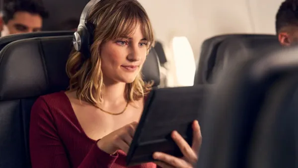 AA Inflight Wifi entertainment tablet use onboard. A woman sitting in an airplane seat, wearing headphones and using a tablet, enjoying in-flight entertainment or working during the flight. She is smiling and appears engaged with her device, with other passengers visible in the background.