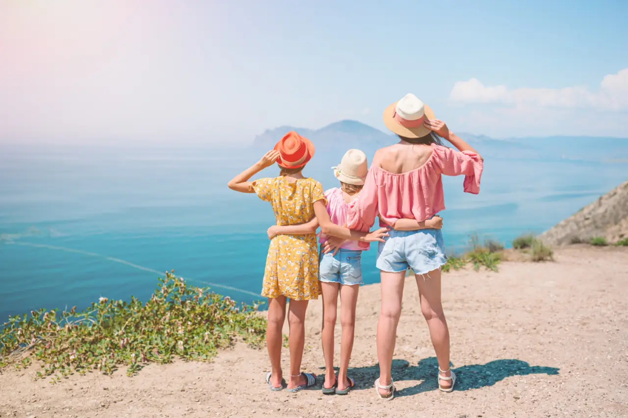 Three people, likely a mother and two children, stand side by side on a cliff overlooking the ocean. They have their backs to the camera and are wearing summer clothing, including sun hats. The scene is bright and sunny, with a clear blue sky and distant mountains visible across the water.