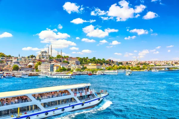 Home 29 A scenic view of the Bosphorus in Istanbul, featuring a large passenger boat in the foreground and the historic skyline with mosques and colorful buildings in the background under a bright blue sky.