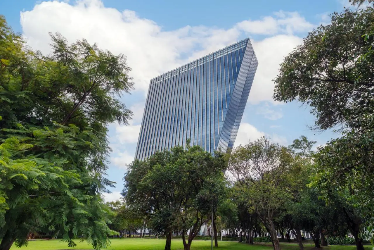 A modern, angular skyscraper with a reflective glass façade, surrounded by trees and greenery, under a partly cloudy sky.