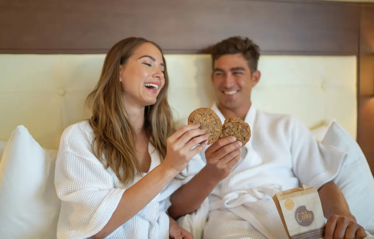 Young couple on a hotel bed wearing white hotel robes enjoying DoubleTree Cookies