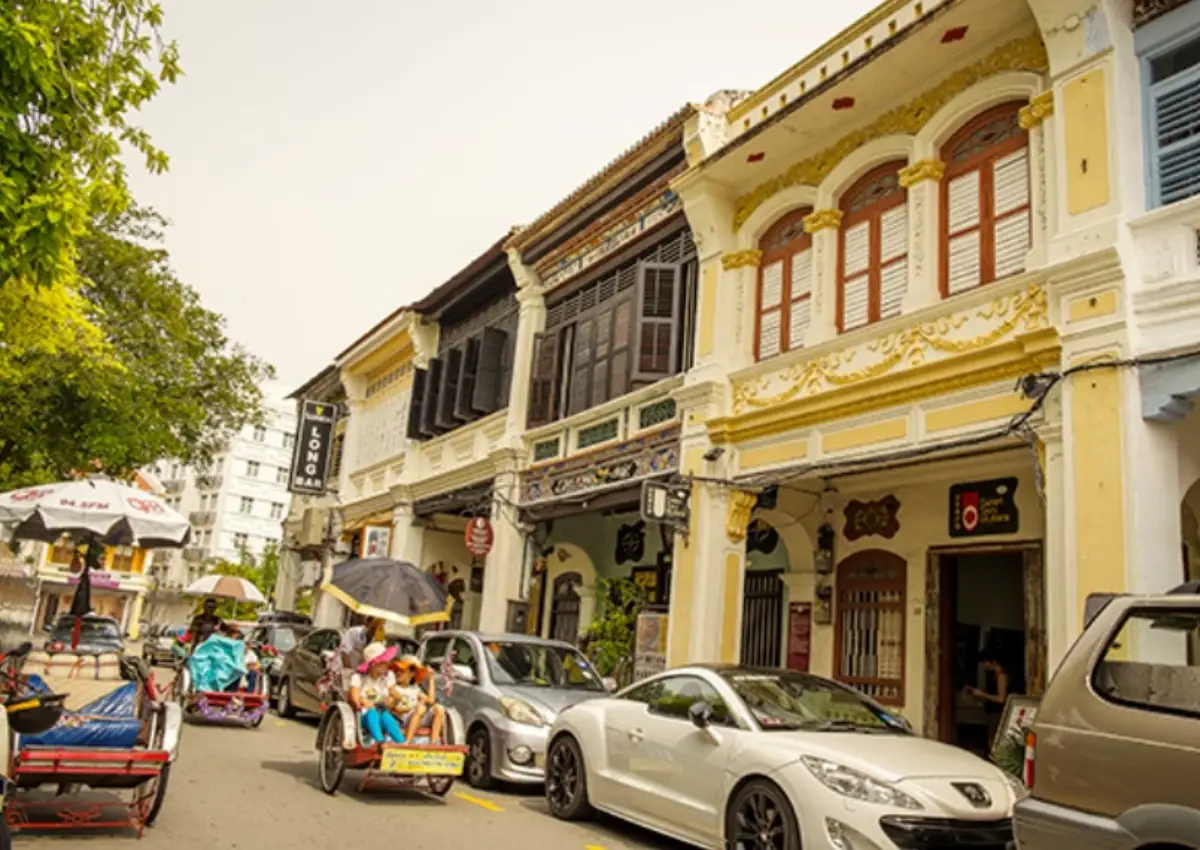 A lively street scene in Georgetown, Penang, showcasing colonial-style shophouses with colorful facades and intricate architectural details. The street is bustling with activity, featuring trishaws carrying tourists, cars, and people walking along the sidewalk under a bright sky.