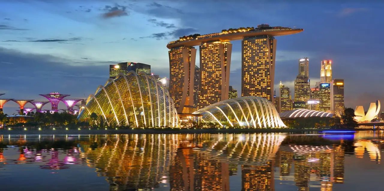 Singapore’s iconic Marina Bay skyline at dusk, featuring the illuminated Marina Bay Sands hotel with its distinctive boat-shaped rooftop and the glass domes of the Gardens by the Bay reflecting in the calm waters.