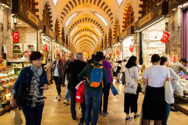 Tourists shopping in the Grand Bazaar, Istanbul, Turkey, walking through a busy, historic market corridor with arched ceilings and shops displaying various goods.