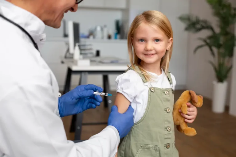 A young girl smiles while receiving a vaccine injection from a healthcare professional, holding a stuffed toy in her other hand.