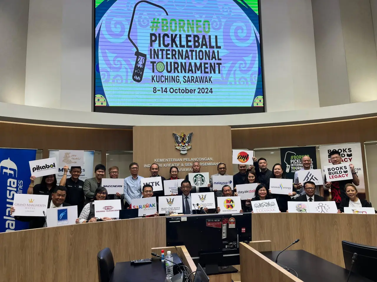 A group of people holding signs representing various sponsors and partners of the Borneo Pickleball International Tournament 2024, standing together in a conference room with a large digital screen in the background displaying the event details.