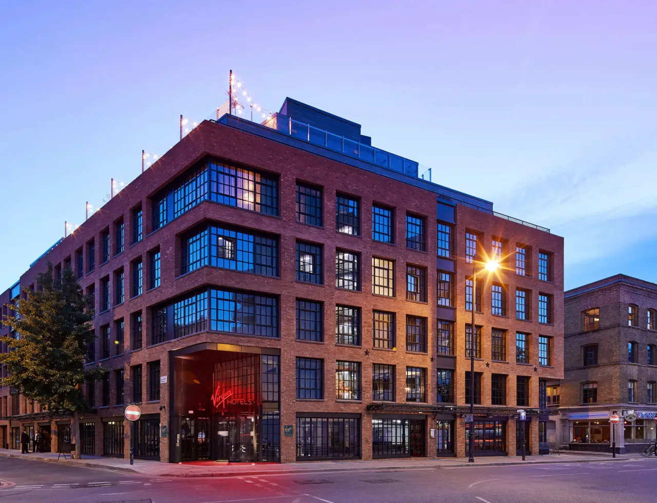 The exterior of Virgin Hotels London-Shoreditch, showcasing a modern brick building with large windows and a red Virgin Hotels sign illuminated at the entrance. The rooftop features string lights, hinting at an inviting rooftop bar area.