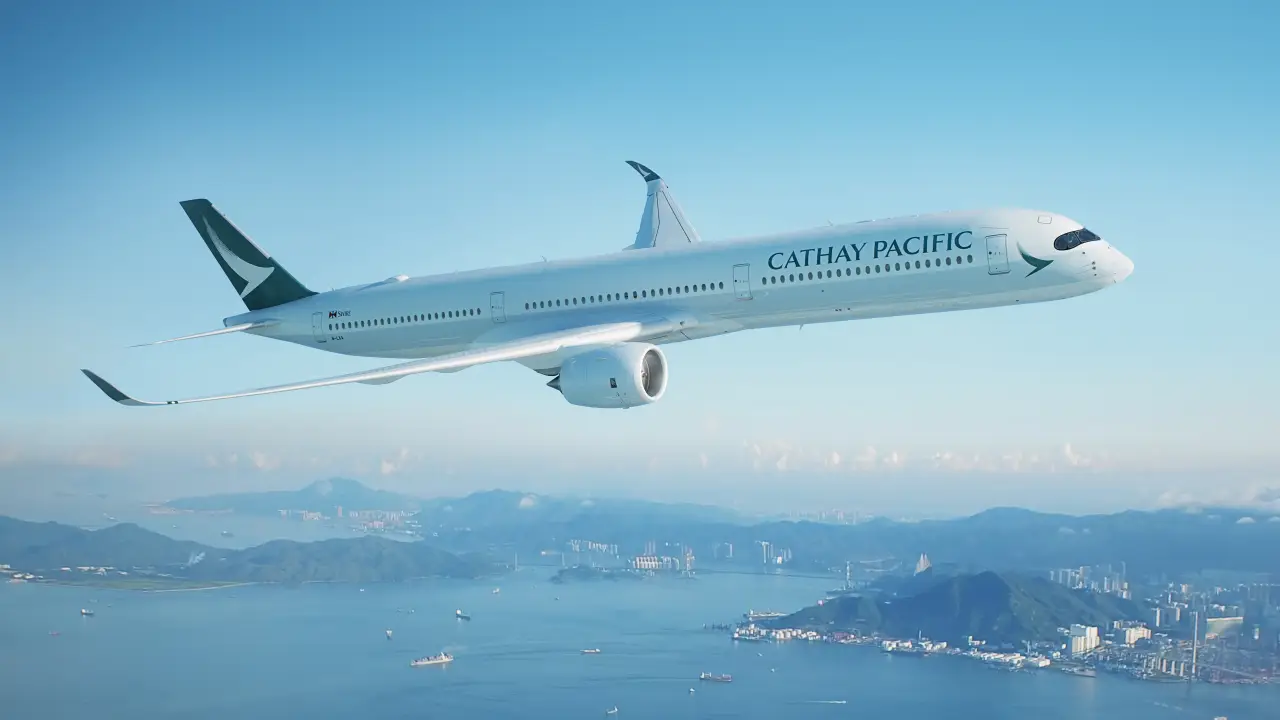 Cathay Pacific Airbus A350 aircraft flying over a coastal landscape with mountains and city skyline in the background.