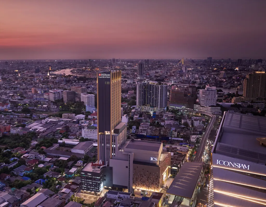 Aerial view of Hilton Garden Inn Bangkok Riverside at sunset, showcasing the tall, modern hotel tower amidst the vibrant cityscape of Bangkok, with the Chao Phraya River and surrounding buildings illuminated in the twilight.