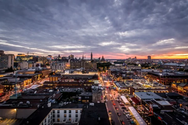 Home 6 A panoramic view of Ottawa’s cityscape at sunset, showcasing a mix of historic and modern buildings with vibrant streets and the distant silhouette of the Canadian Parliament buildings.