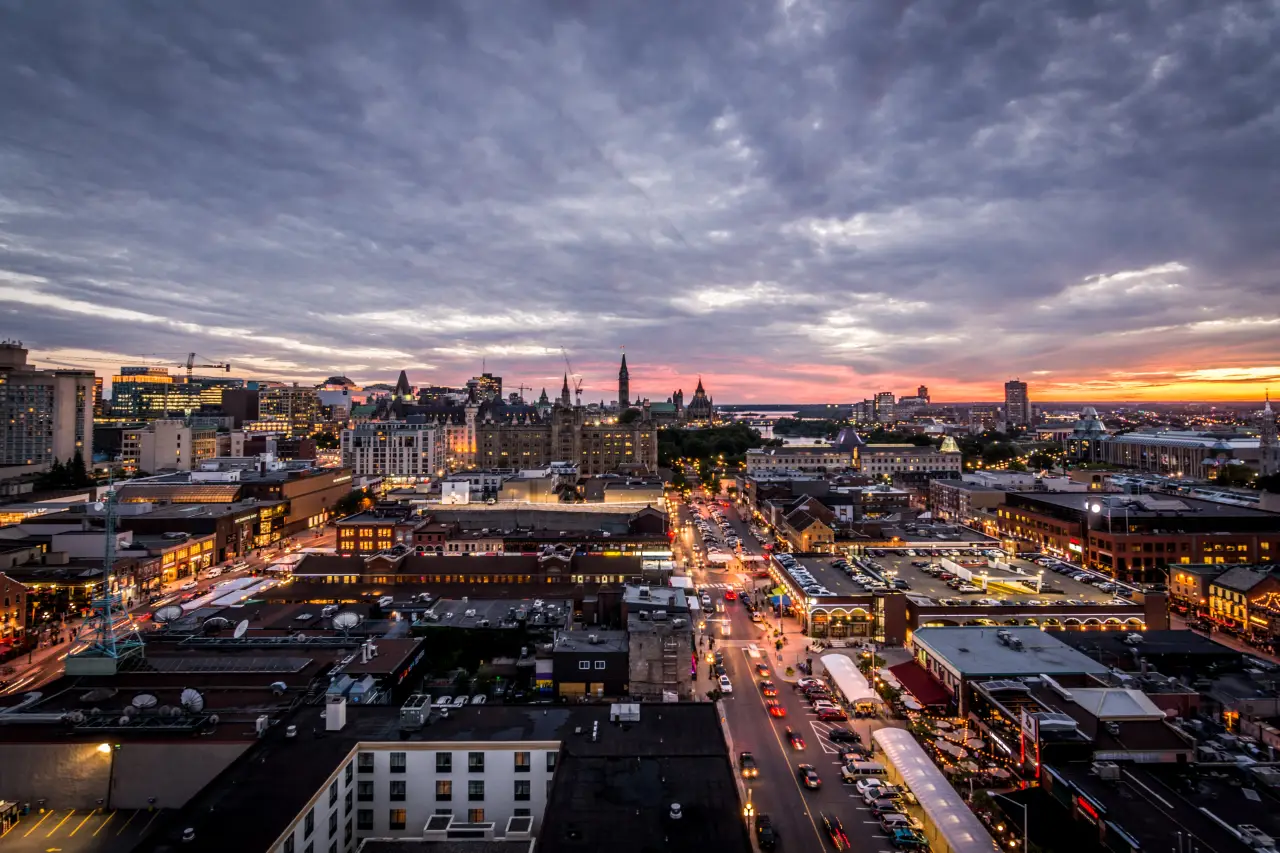 A panoramic view of Ottawa’s cityscape at sunset, showcasing a mix of historic and modern buildings with vibrant streets and the distant silhouette of the Canadian Parliament buildings.