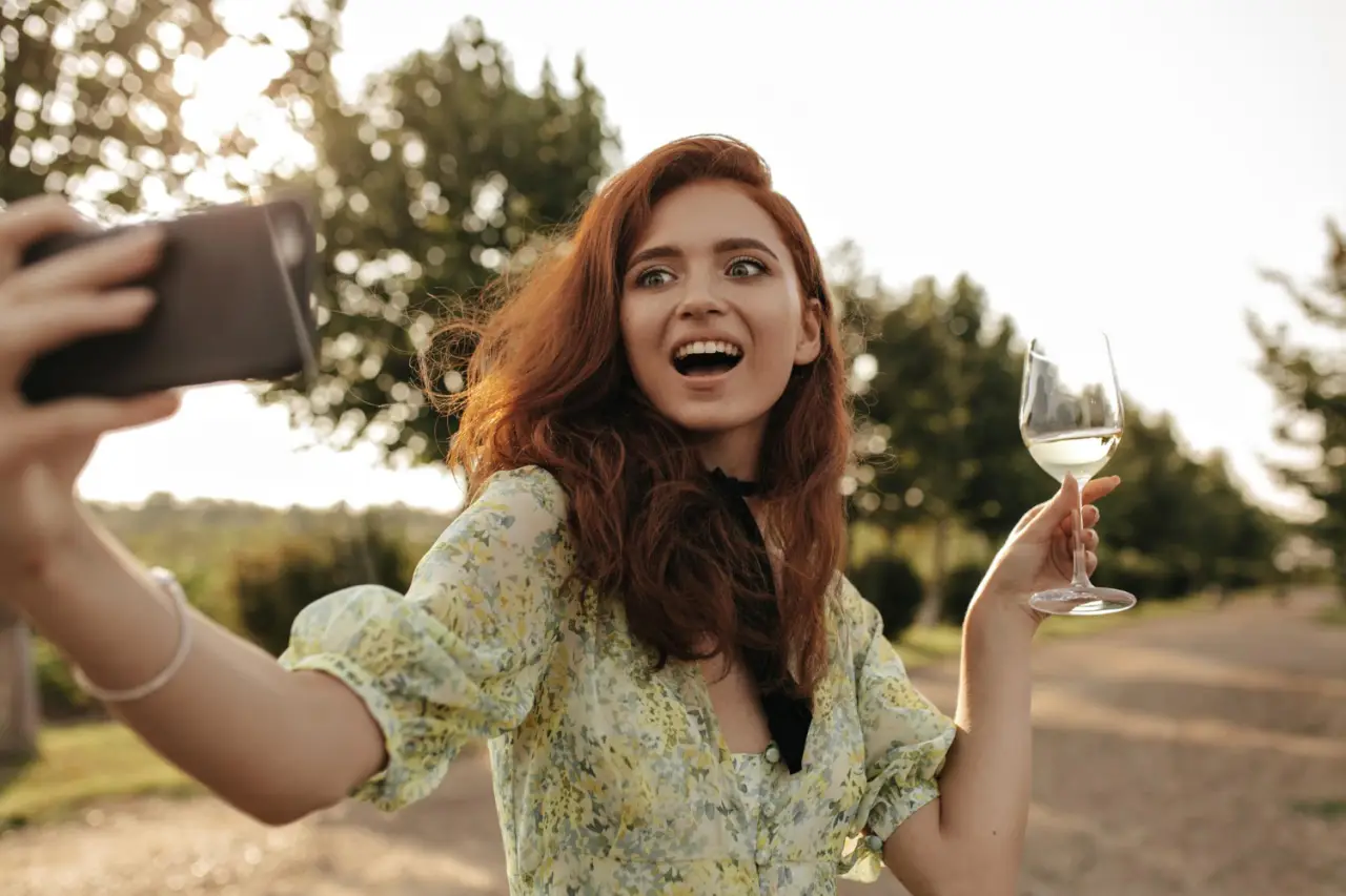 A young woman with red hair, wearing a floral dress, takes a selfie while holding a glass of white wine outdoors, with a backdrop of greenery and sunlight filtering through the trees.