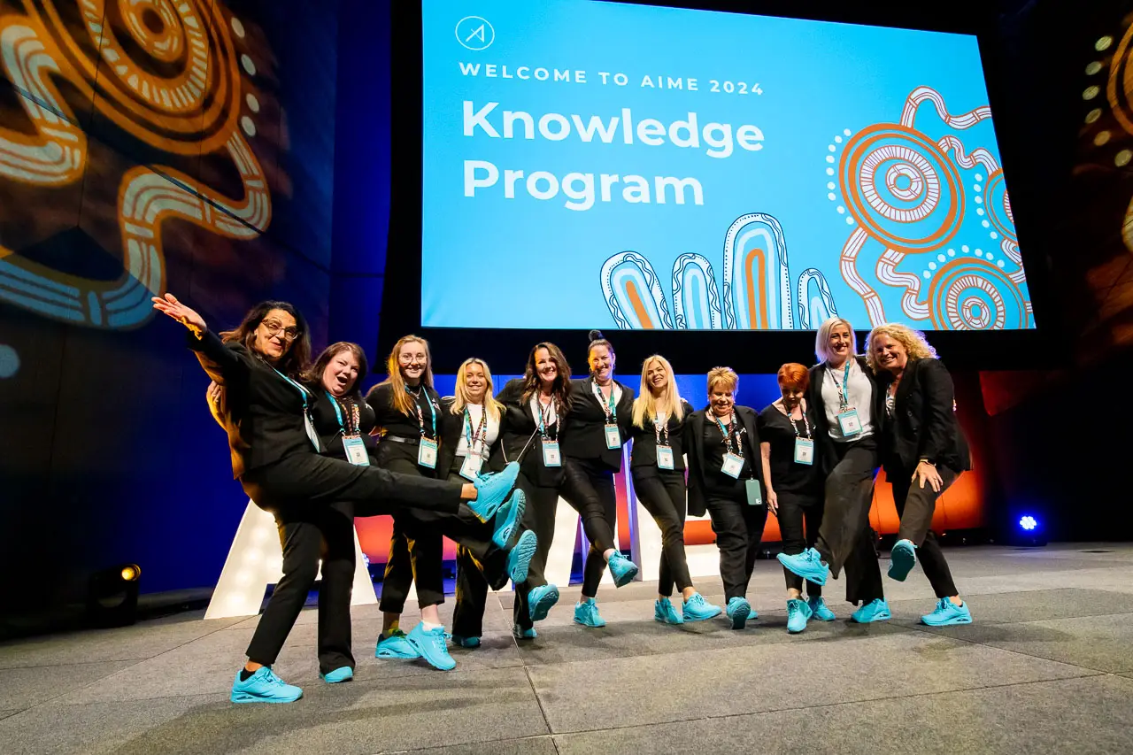 Group of event professionals at AIME 2024 Knowledge Program posing on stage with matching blue sneakers