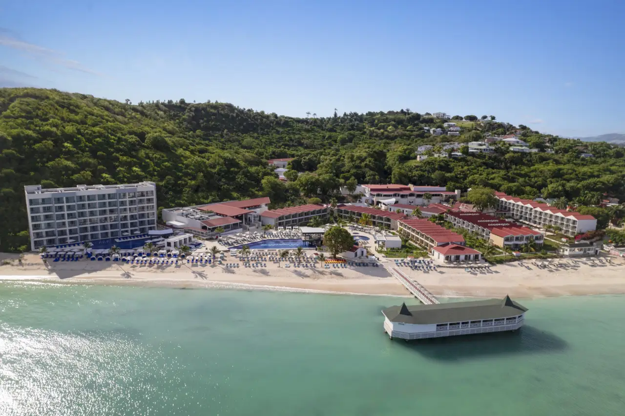 Aerial view of Royalton CHIC Antigua resort featuring modern buildings, a beachfront pool area, and a white-sand beach surrounded by lush green hills.