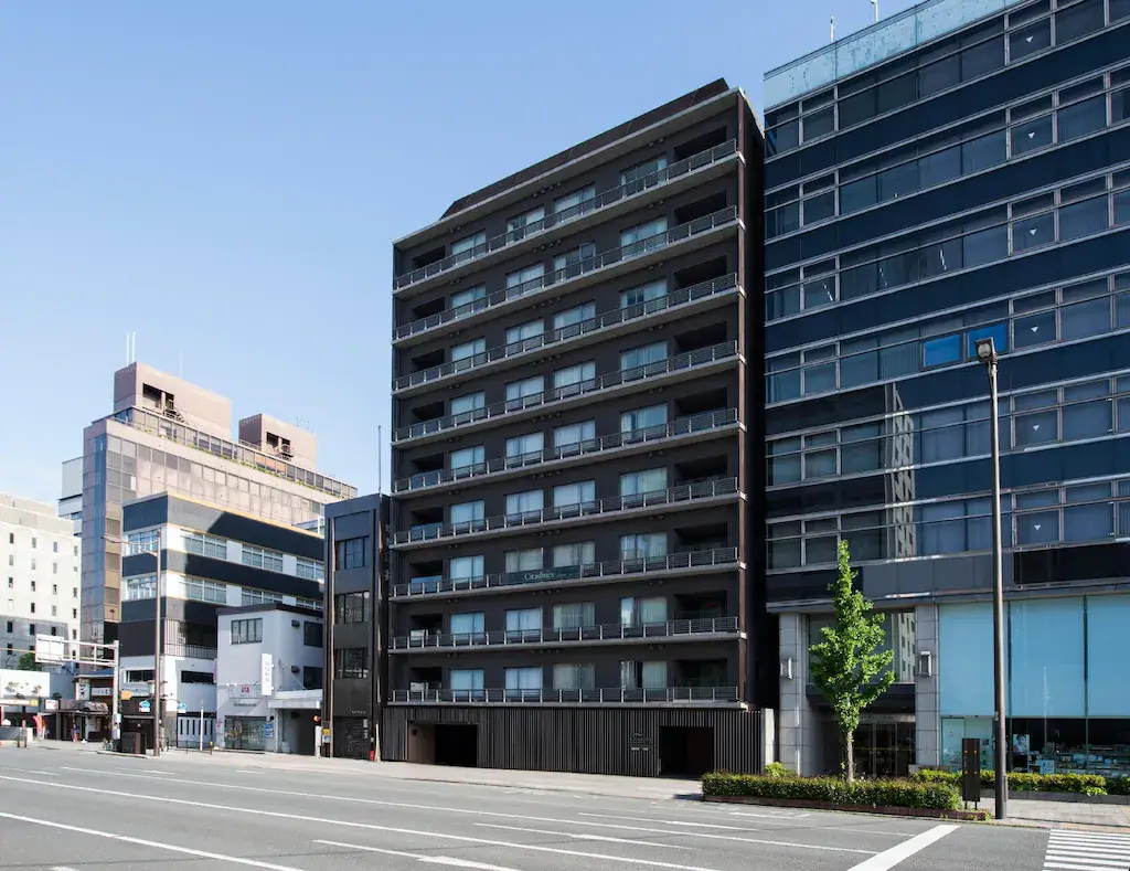 Capitaland Ascott Trust Divests Citadines Karasuma-Gojo Kyoto 7 Exterior view of Citadines Karasuma-Gojo Kyoto, a modern multi-story residential building in Japan. The property features a sleek black facade with balconies on each floor, situated on a bustling street lined with other contemporary buildings.