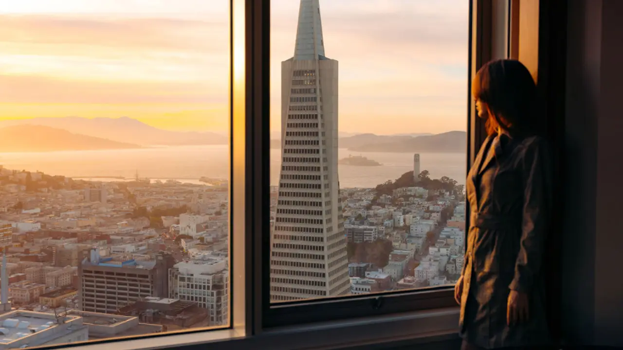 Discover What Four Seasons Guests Are Driving Next 16 Woman looking out the window of Four Seasons Hotel San Francisco at Embarcadero with a stunning view of the city skyline, including the Transamerica Pyramid and bay.