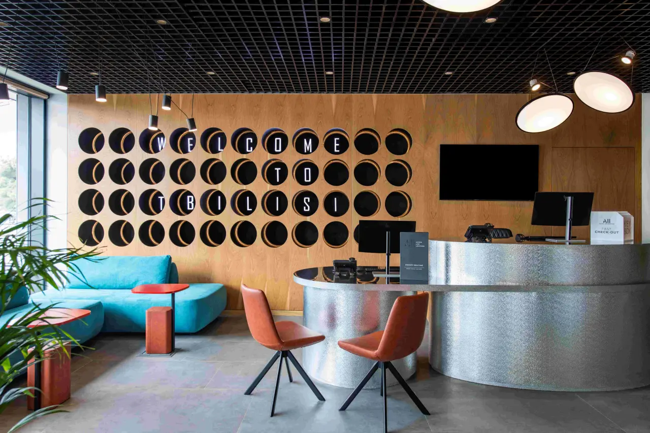 Modern reception area at ibis Tbilisi Airport hotel featuring a stylish circular wall design that spells “Welcome to Tbilisi” in illuminated letters. The lobby is decorated with a mix of contemporary furniture, including turquoise seating and red chairs, and has a sleek, metallic front desk.