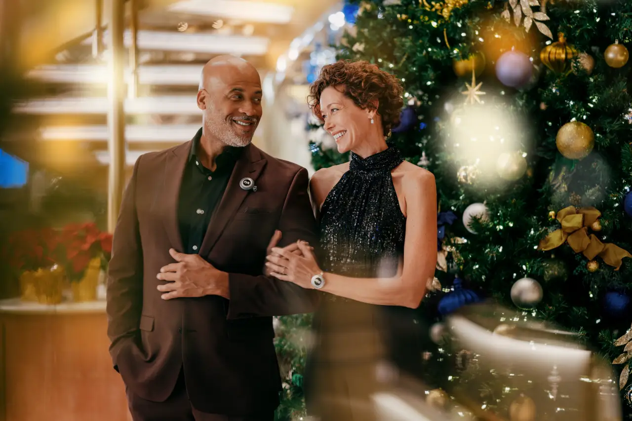 A couple dressed elegantly, standing in front of a decorated Christmas tree aboard the Sun Princess cruise ship.