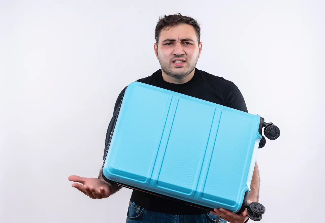 A confused young man in a black t-shirt holding a blue suitcase, expressing frustration or uncertainty, standing against a white background.