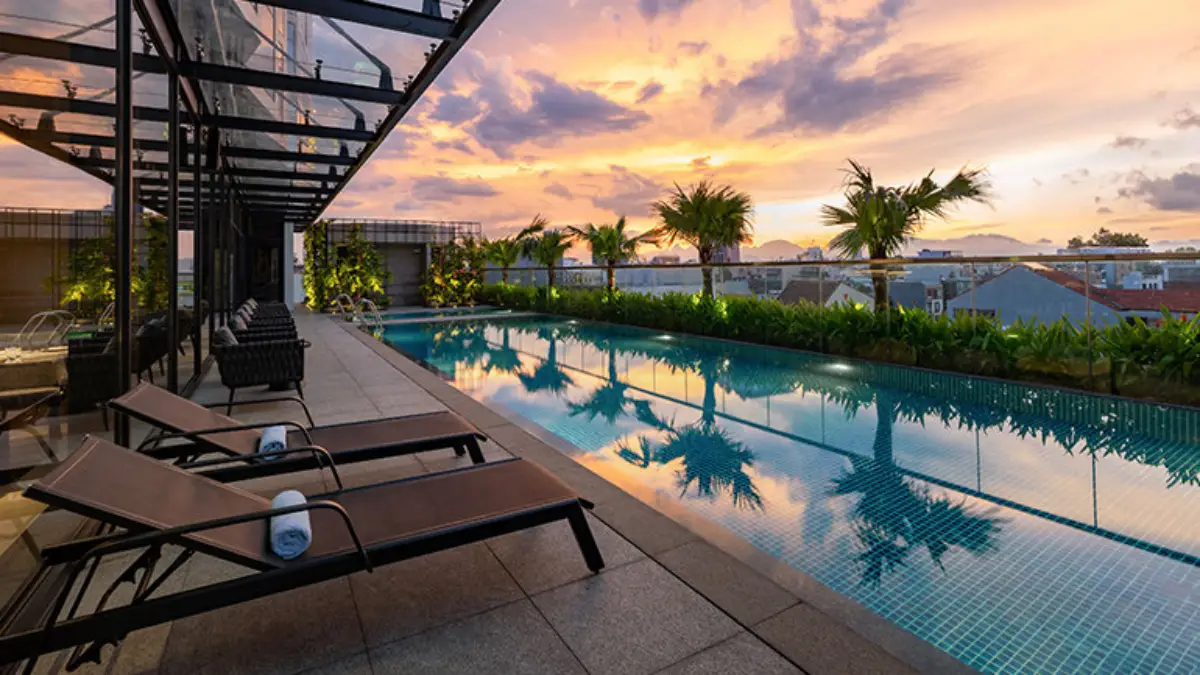 Crowne Plaza Danang City Centre's rooftop swimming pool at sunset with lounge chairs and palm trees, offering a view of Danang cityscape.