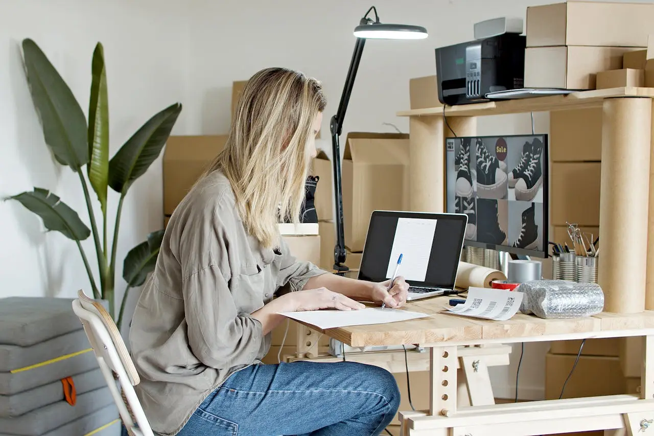 A woman working at a desk in a home office, surrounded by cardboard boxes, using a laptop and taking notes.