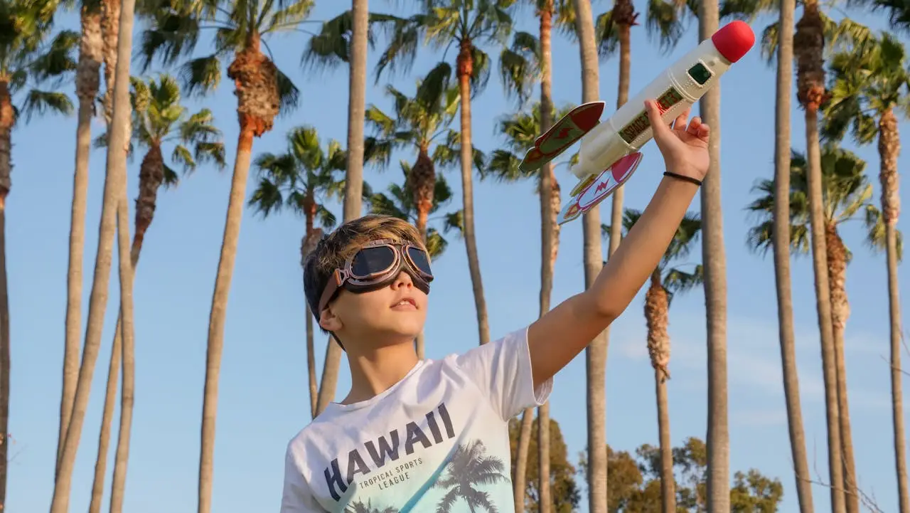 A young boy wearing aviator goggles and a white “Hawaii” t-shirt holds a toy rocket against a backdrop of tall palm trees and a clear blue sky.