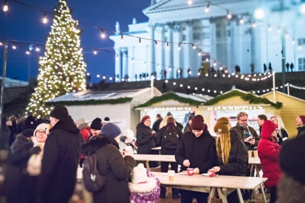People enjoying festive food and drinks at Helsinki’s Christmas market in Senate Square, with a decorated Christmas tree and Helsinki Cathedral in the background. Credit: Jussi Hellsten