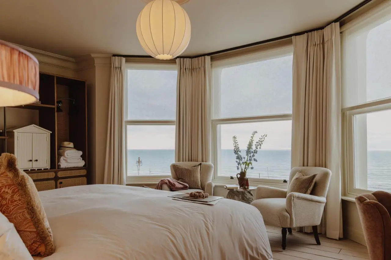 A cozy and elegant hotel room at No. 124 Brighton, featuring large bay windows with a stunning view of the sea, soft beige curtains, and warm, inviting decor.