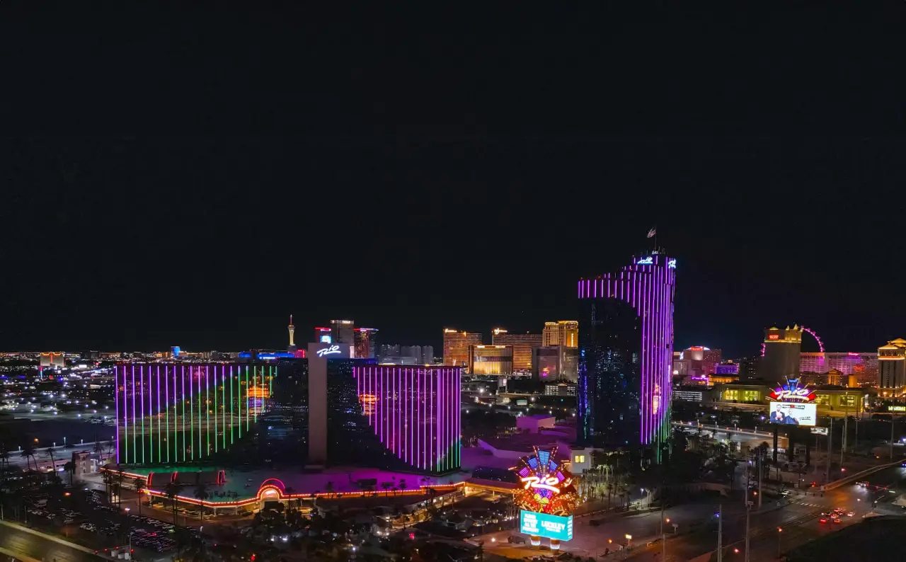 Nighttime view of the Rio Hotel and Casino in Las Vegas, illuminated with vibrant purple and green lights, with the Las Vegas Strip in the background.