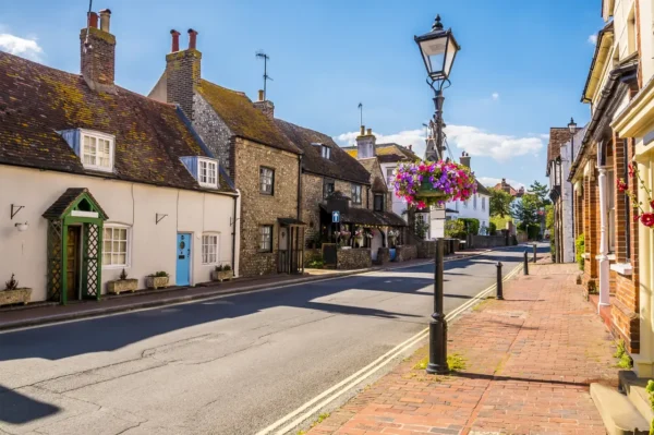 A charming street in Rottingdean, East Sussex, featuring historic cottages, vibrant hanging flower baskets, and a sunny blue sky.