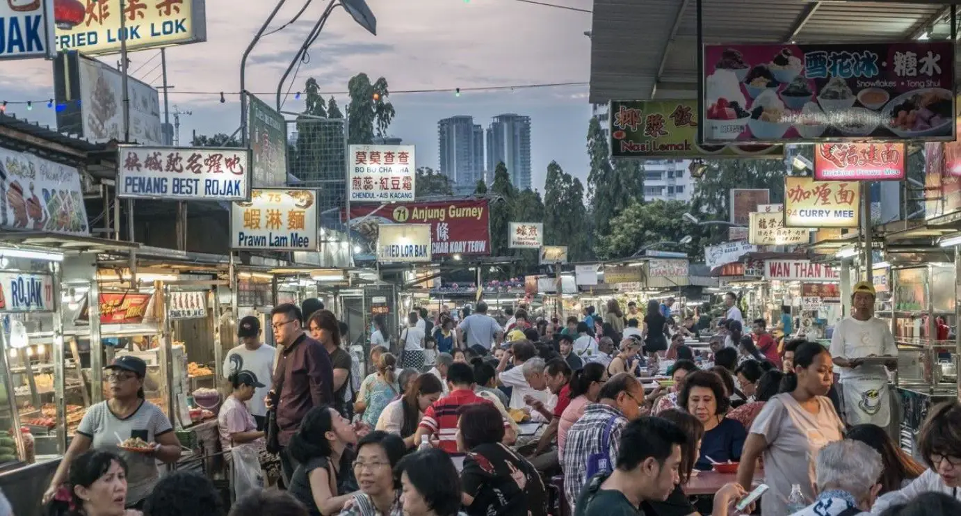 A bustling Penang street food market at dusk, filled with locals and tourists enjoying dishes under brightly lit food stalls and colorful signboards.