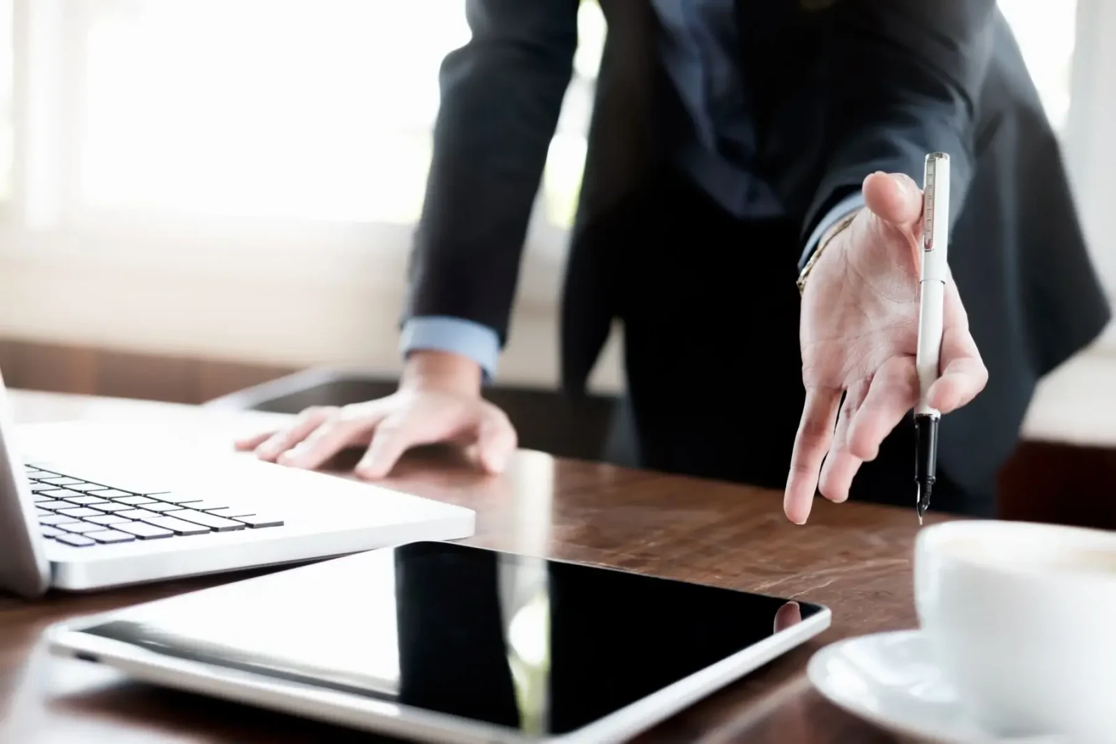 A professional hand holding a pen, gesturing towards a wooden desk with a laptop, a tablet, and a cup of coffee.