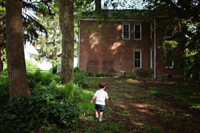 A young child walking alone in a backyard surrounded by trees and greenery.