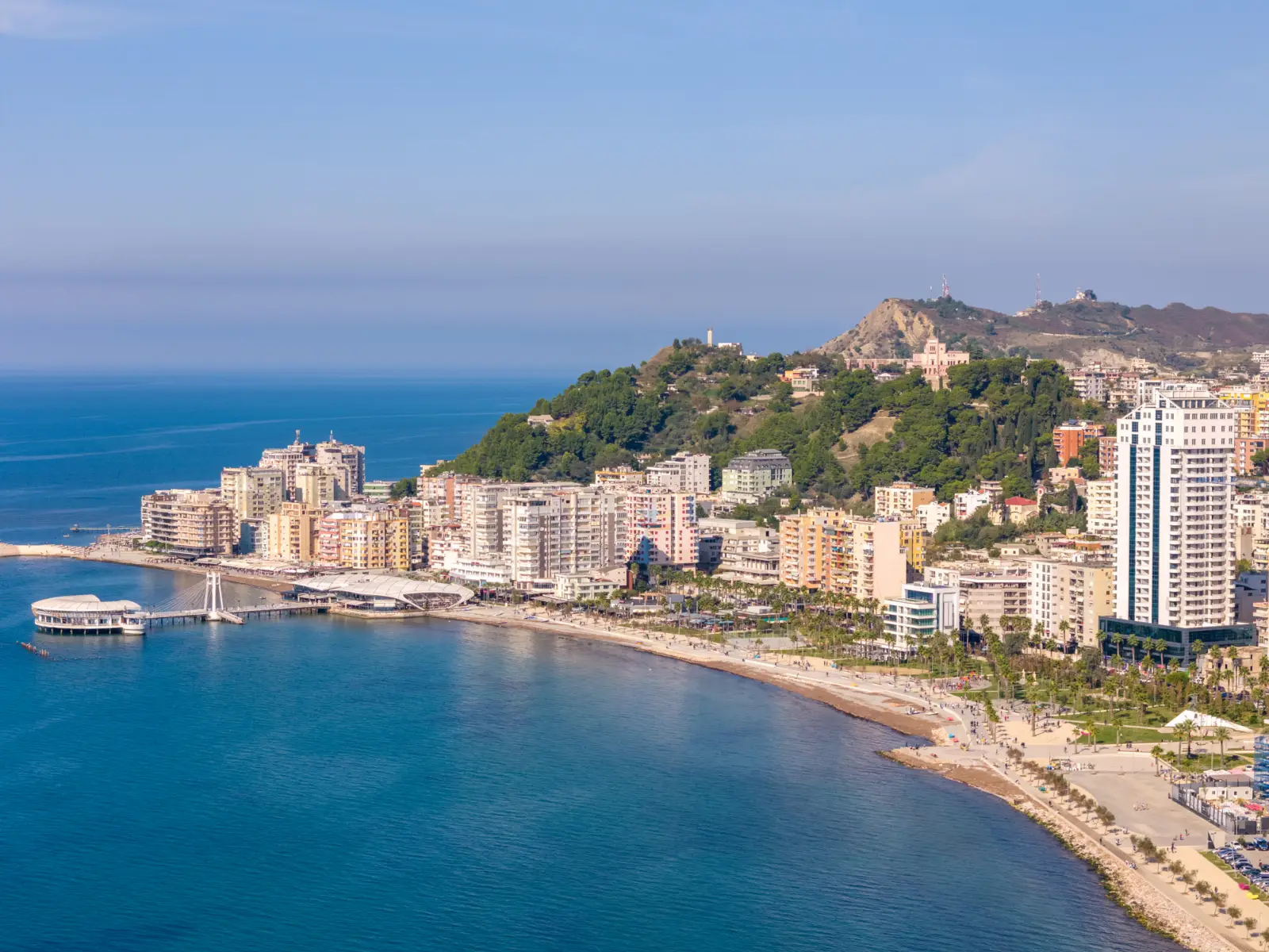 A vibrant coastal view of Durres, Albania, one of the cheapest Mediterranean cities, featuring modern buildings, a scenic pier, sandy beaches, and lush hills.