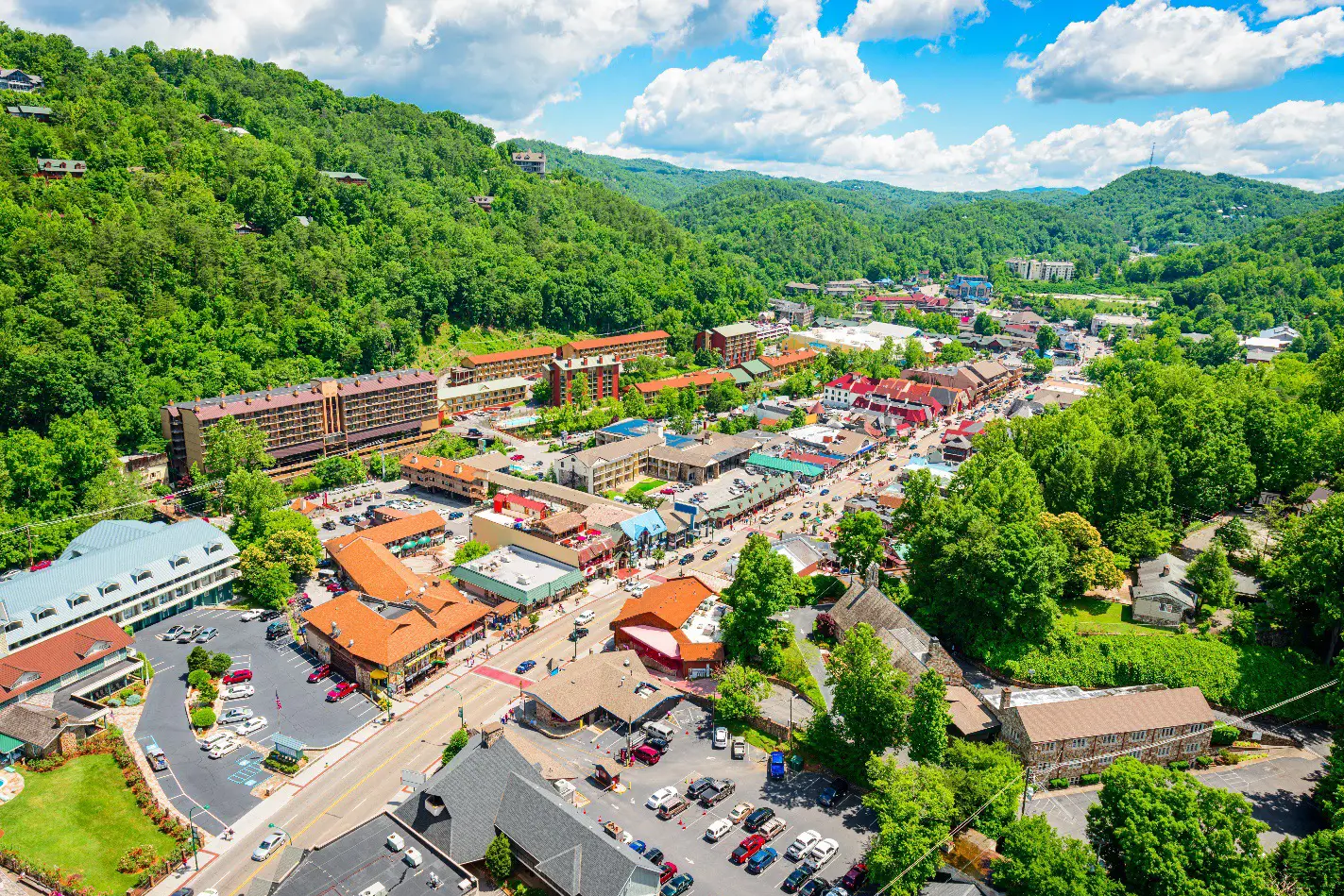 A scenic aerial view of downtown Gatlinburg, Tennessee, surrounded by lush green hills under a bright blue sky with scattered clouds.