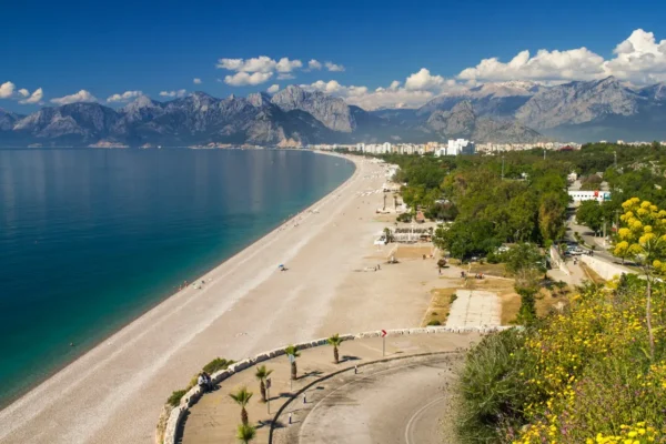 A panoramic view of Konyaaltı Beach in Antalya, Turkey, with its expansive shoreline, crystal-clear waters, lush greenery, and the dramatic Taurus Mountains in the background.