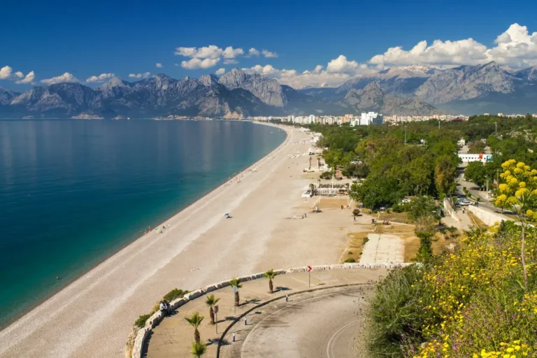 A panoramic view of Konyaaltı Beach in Antalya, Turkey, with its expansive shoreline, crystal-clear waters, lush greenery, and the dramatic Taurus Mountains in the background.