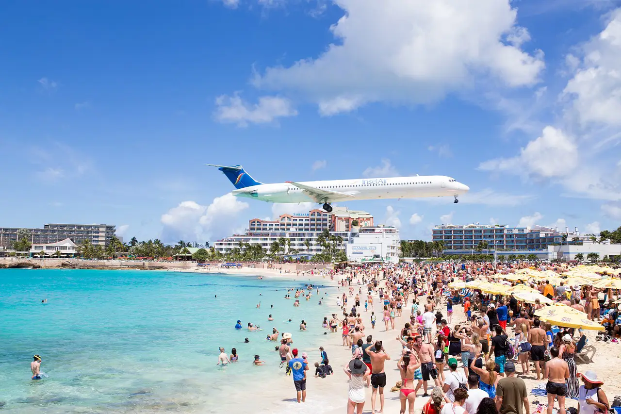 A low-flying airplane approaches Princess Juliana International Airport, soaring above a crowded Maho Beach with turquoise waters and sunbathing tourists.