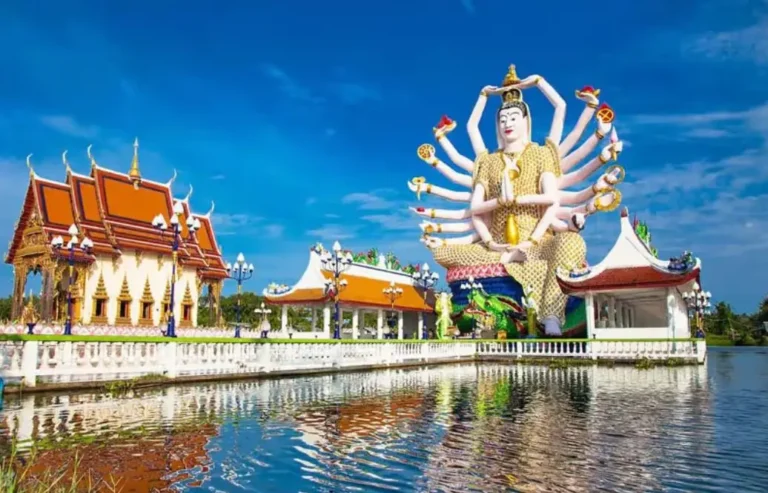 A stunning temple in Thailand with a giant multi-armed statue of Guanyin reflected in a serene pond under a bright blue sky.