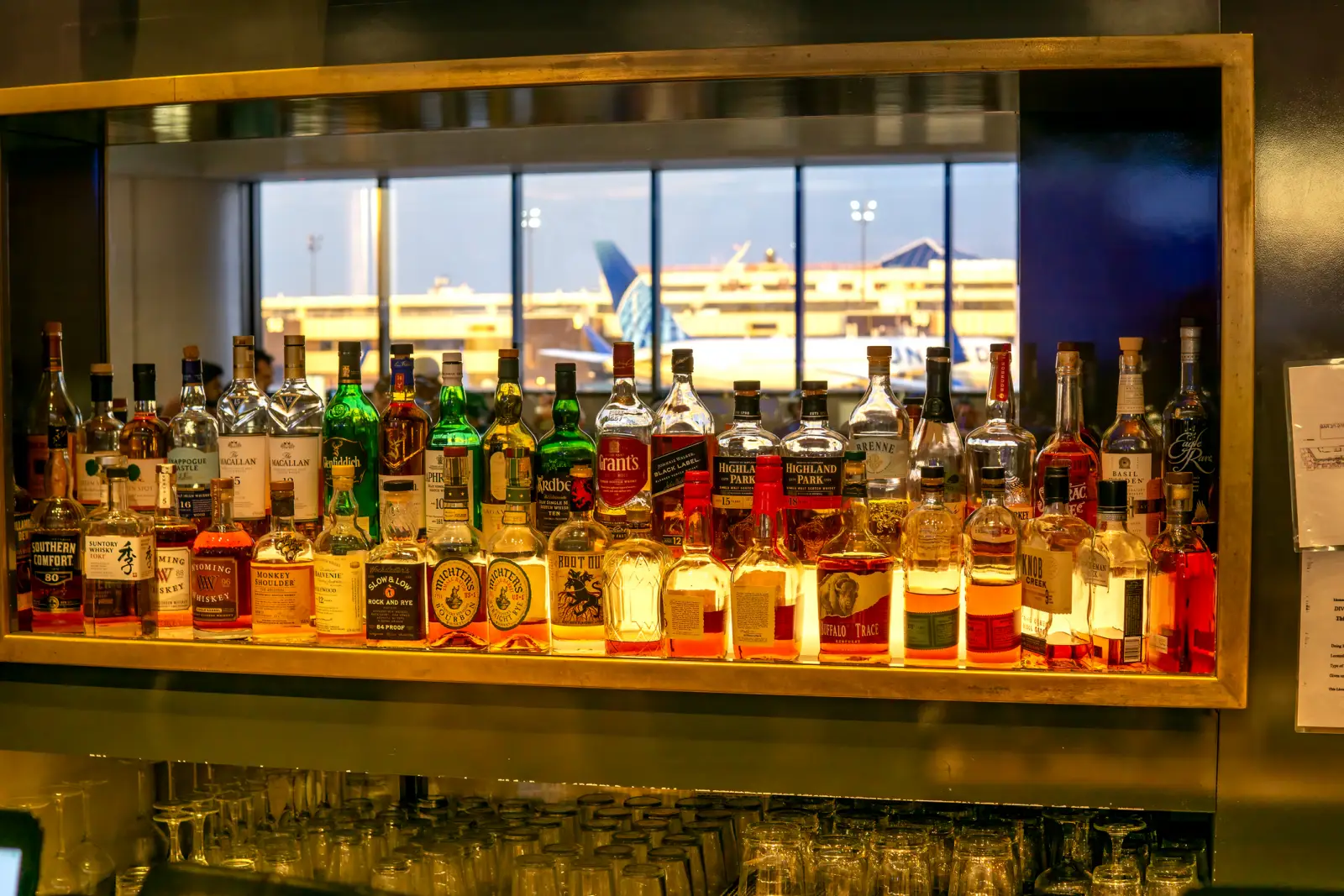 A variety of liquor bottles displayed on a shelf in an airport bar, with an airplane visible through the window in the background.