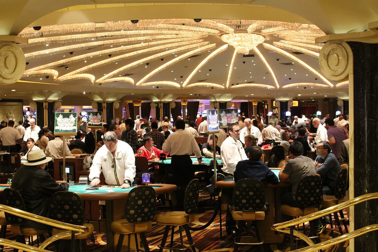 A lively casino in Las Vegas with poker tables, dealers, and players under a grand chandelier.