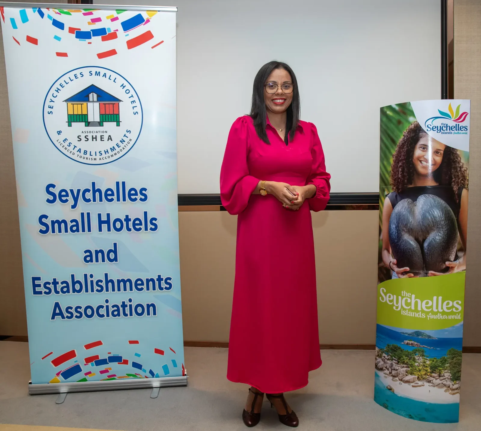 Sherin Francis, wearing a bright pink dress, stands smiling between two Seychelles Tourism banners.