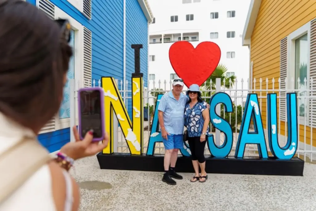 Two tourists pose in front of a colorful “I ♥ NASSAU” sign as another person takes their photo.